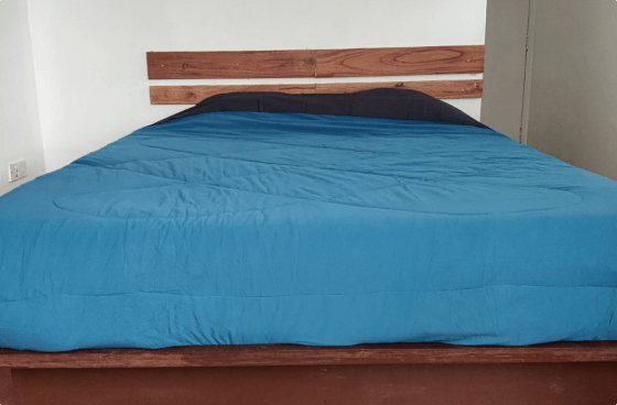 Bedroom with a simple design featuring a wooden bed frame, blue comforter, and a rustic wooden headboard against a plain white wall.